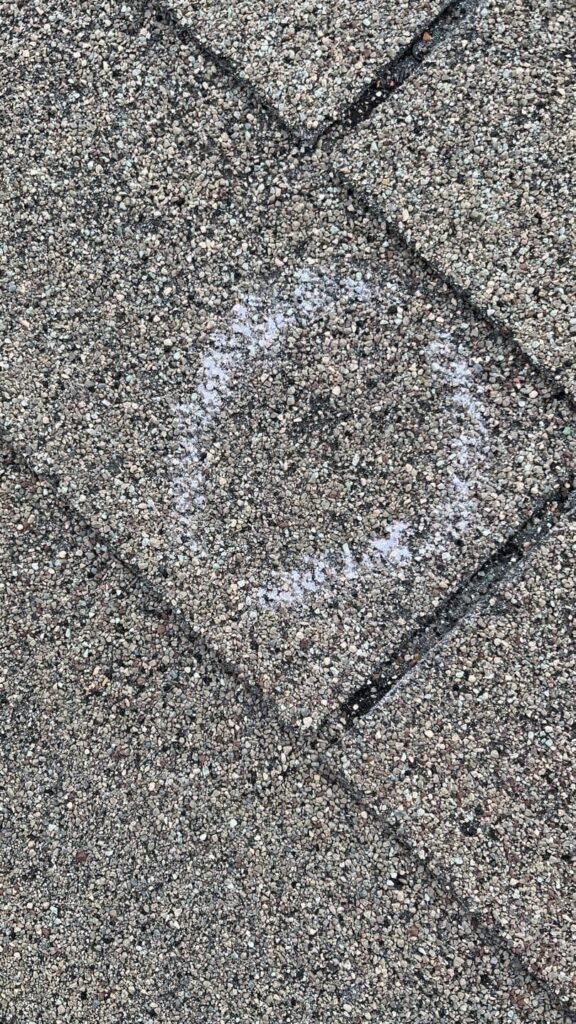 A photo of a roof with hail damage. The roof is covered in small dents and pockmarks, and there are several large holes in the shingles. The gutters are also damaged, and there is some debris scattered around the ground.